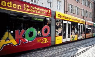 Seitlich vollbeklebte Straßenbahn mit bunter Werbegestaltung für Kindermode. Verkehrsmittelwerbung wird hier als mobile Markenbotschaft im Stadtbild eingesetzt.