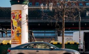 Litfaßsäule an einer urbanen Allgemeinstelle mit auffälliger Plakatwerbung – im Hintergrund ein Bahnhof und vorbeifahrende Fahrzeuge, im Vordergrund ein geparktes Auto