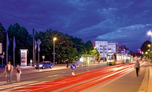 Nächtliche Szene an einer Haltestelle mit Lichtspuren vorbeifahrender Autos. Leuchtende Ganzsäule an strategischer Ganzstelle sorgt auch abends für hohe Sichtbarkeit