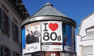 Klassische Litfaßsäule mit Plakaten zur 80er-Jahre-Musik vor Altbaufassaden – typische Werbefläche an einer zentralen Allgemeinstelle.