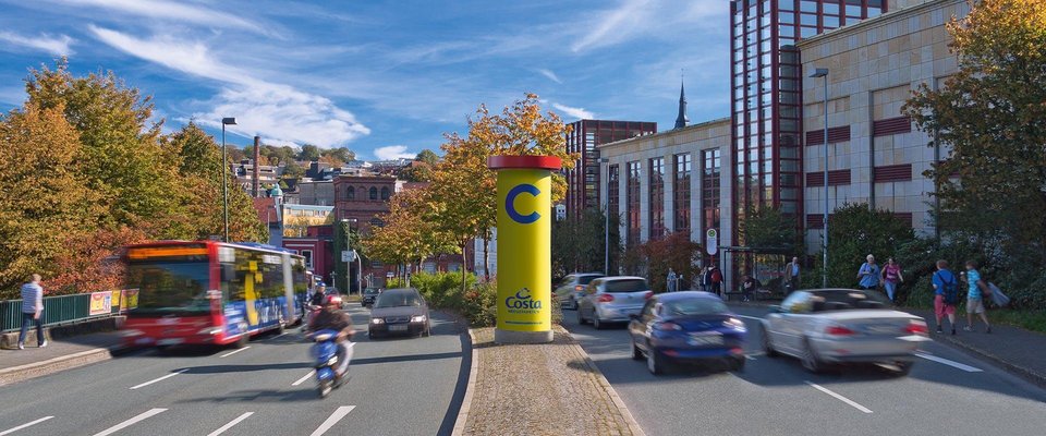 Zentrale Litfaßsäule an einer stark frequentierten Allgemeinstelle zwischen Fahrbahnen mit fließendem Verkehr, Fußgängern und einem vorbeifahrenden Stadtbus – ideal platzierte Werbefläche in urbanem Umfeld.